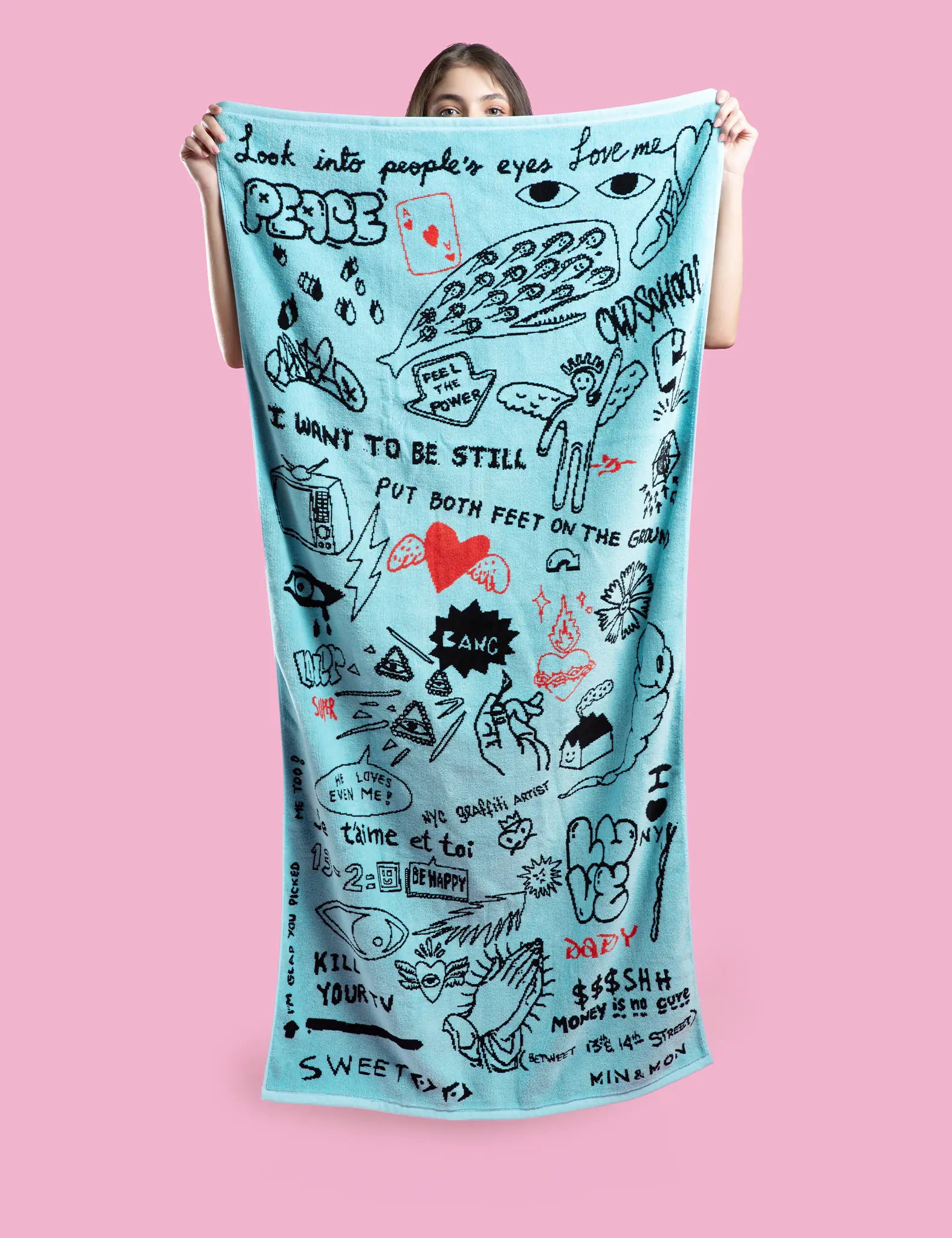 Woman holds up a light blue towel covered in bold, doodle-style black and red graphics and phrases like “Look into people’s eyes,” “LOVE ME,” and “PUT BOTH FEET ON THE GROUND.” Pink background.