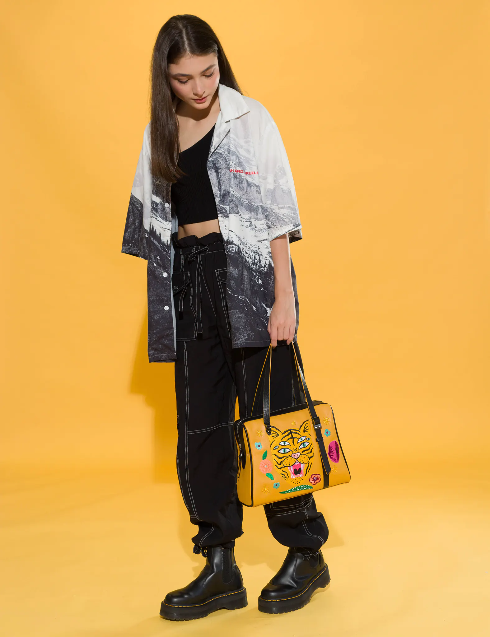 Woman in black cargo pants, platform boots, and a monochrome printed shirt holds a mustard yellow handbag embroidered with a three-eyed tiger and colorful flowers. Yellow backdrop, edgy style.