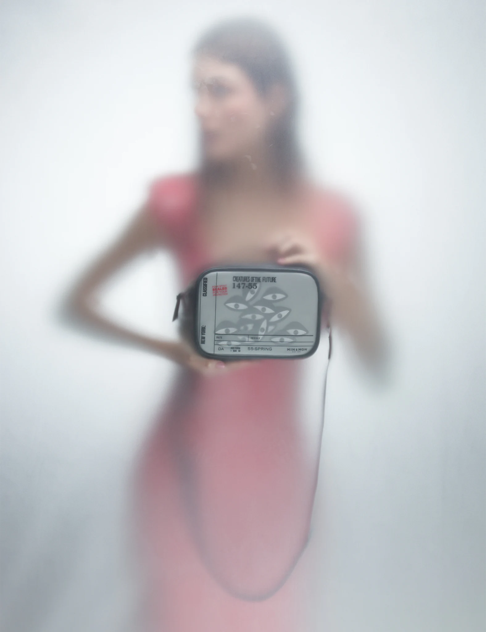 A woman in a coral dress is seen through a foggy surface, holding a Min & Mon crossbody bag featuring a bold graphic of multiple eyes and futuristic typography.