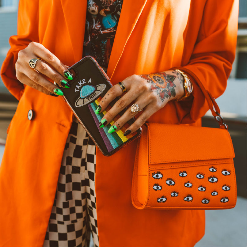Close-up of tattooed hands with green alien nail art holding a “Take a Trip” rainbow wallet and bright orange handbag with embroidered eyes. Person wears an orange blazer and checkered pants.