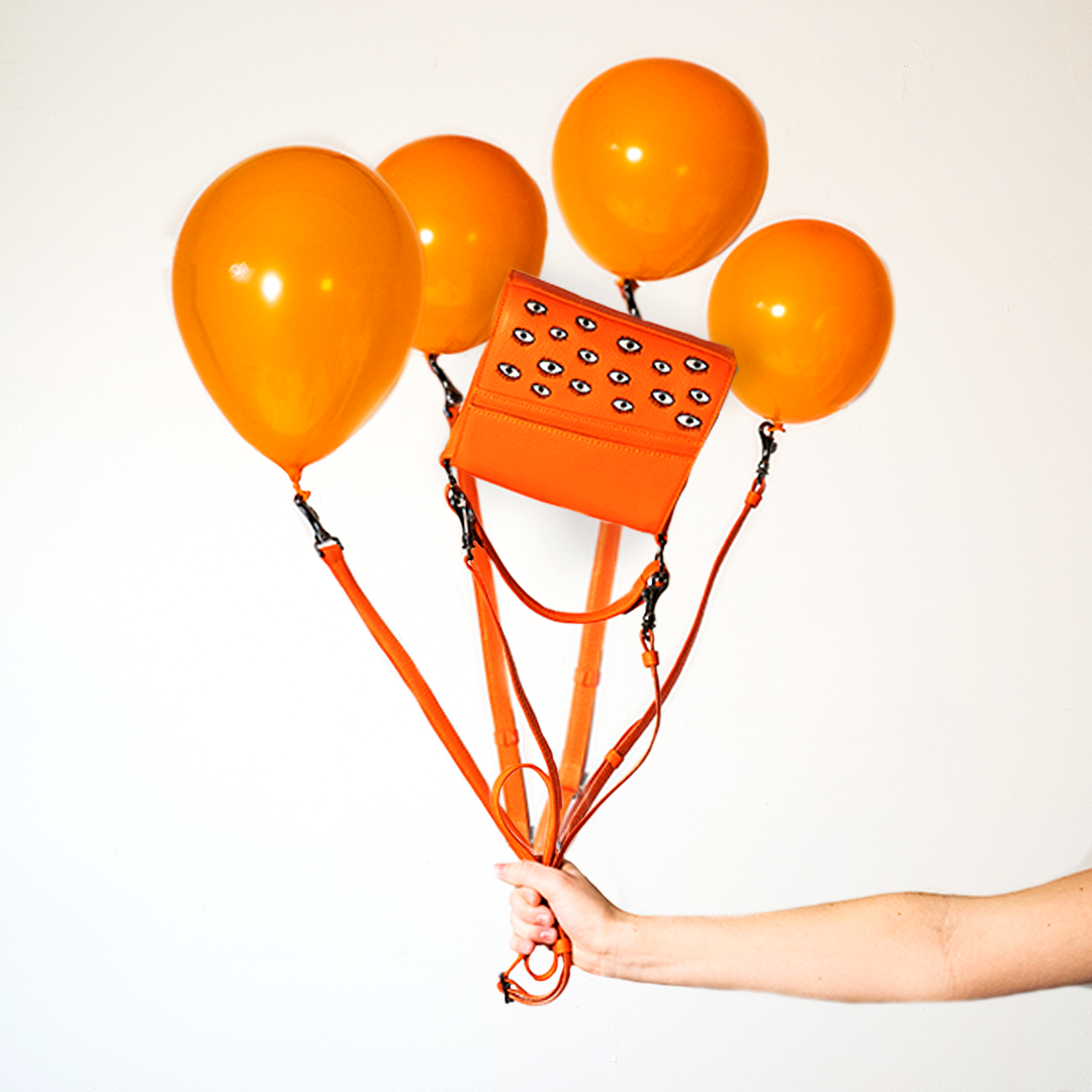 Hand holds four orange balloons, each attached to an orange handbag decorated with embroidered eyes, creating the illusion that the bag is floating. Set against a plain white background.